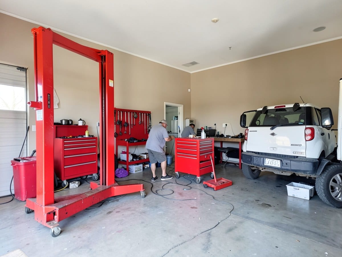 spacious and well-lit garage, with a red hydraulic lift, beige wall, red tool chest, and red tool rack, houses a white car, truck, and red toolbox.