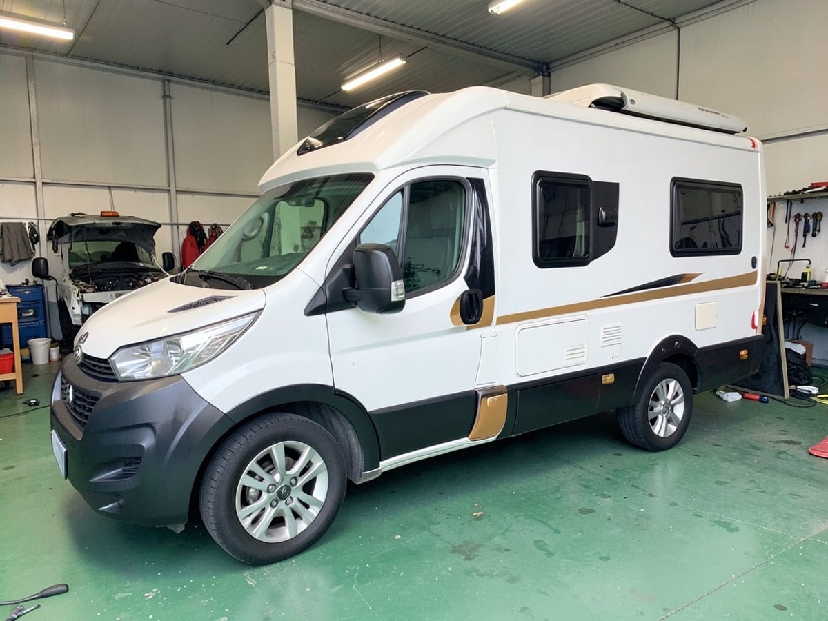 white and black recreational vehicle with tan accents is parked on a green surface, featuring a sunroof, black kitchenette, and silver-toned wheels.
