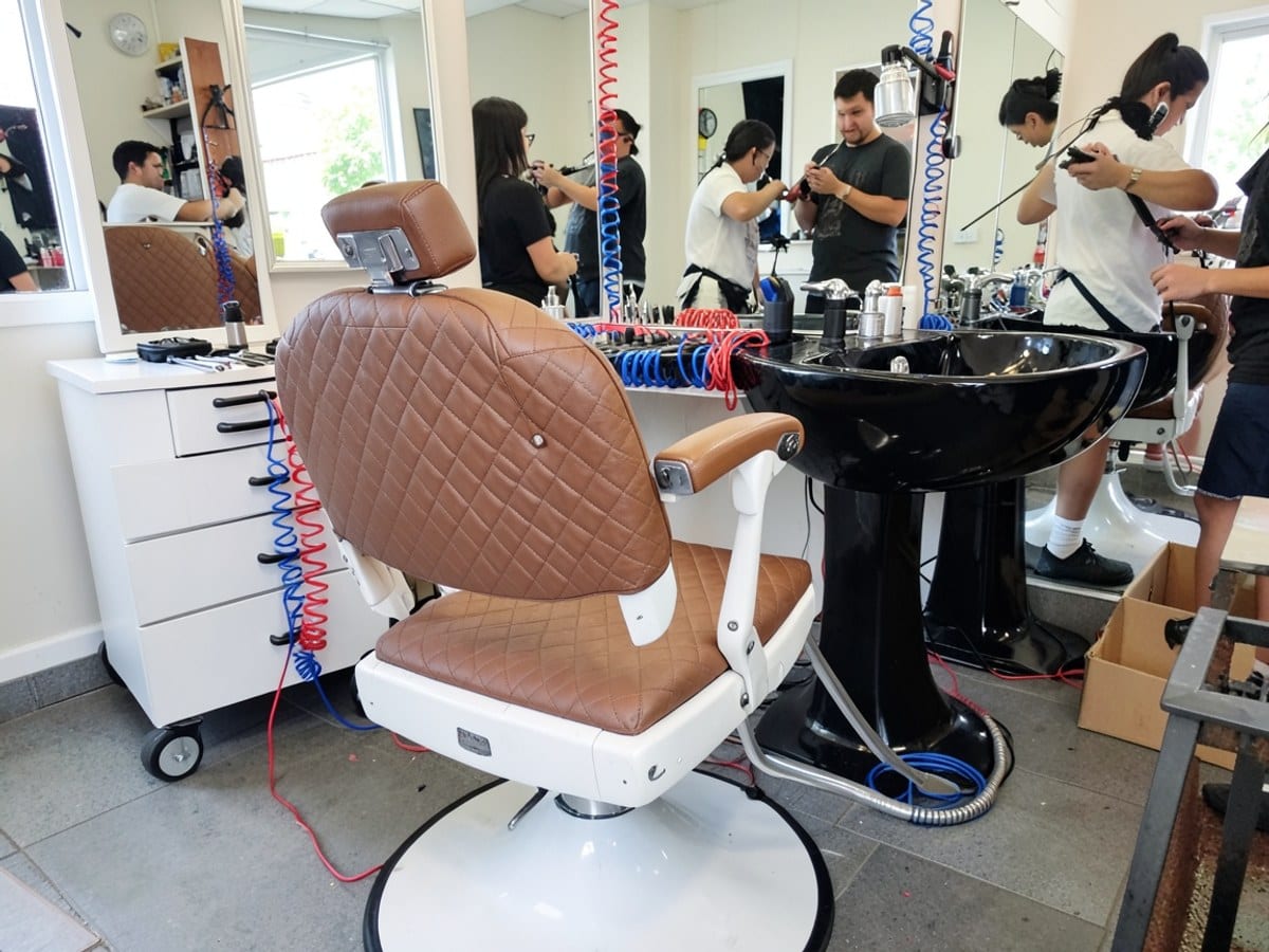 well-equipped barber's chair with a white base and brown quilted fabric is accompanied by a white tool chest and a black sink, all adorned with mirrors and accented by red and blue coiled cords.
