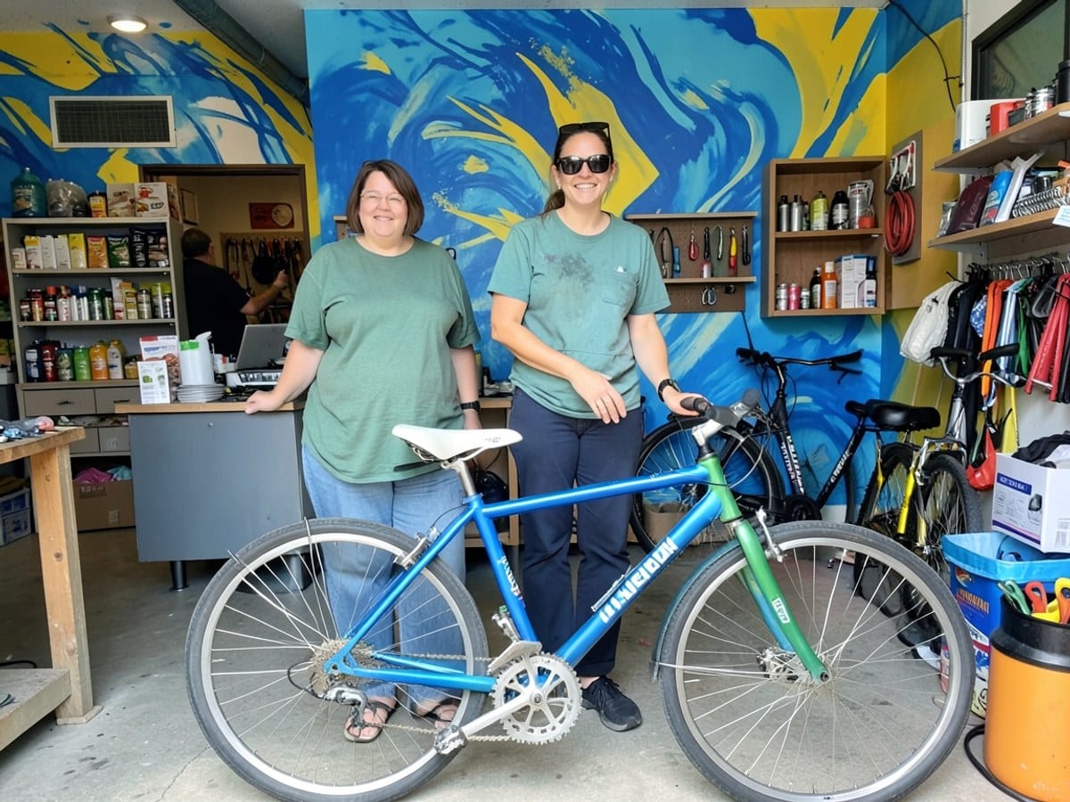 woman in green and sunglasses stands next to a blue-green bicycle with a white seat and handlebars, against a vibrant blue and yellow mural backdrop.