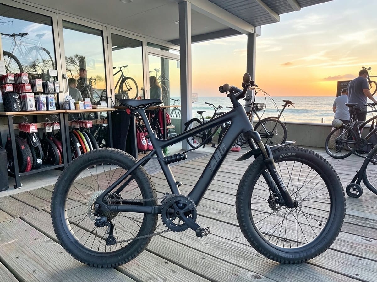 black TST Fat Tire E-Bike with dual suspension, 20" x 4" fat tires, and 7-speed gears is parked on a wooden boardwalk, with the ocean and sunset in the background.