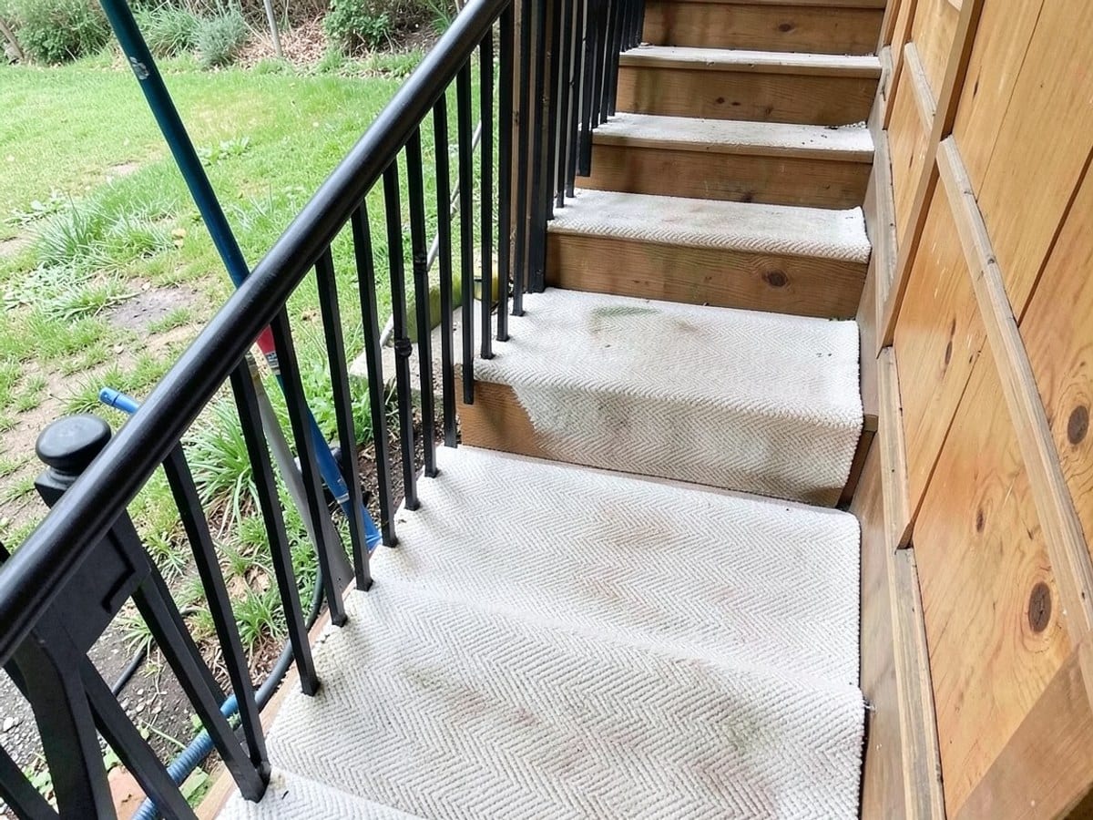 wooden staircase with a white herringbone-patterned runner ascends alongside a black railing, backed by a grassy area and wooden panels.