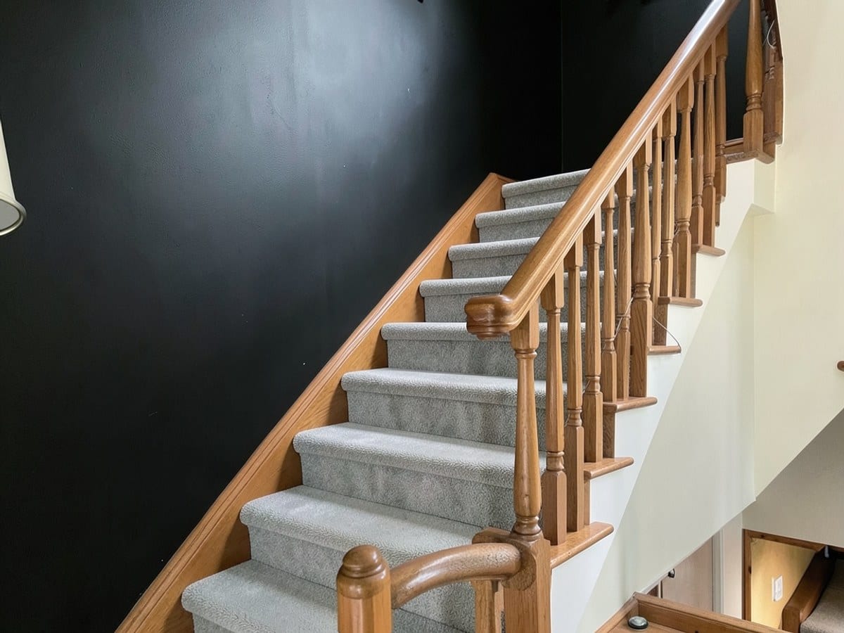 wooden staircase with a wooden banister and light gray carpeted steps ascends towards a black wall, creating an elegant and visually appealing composition.