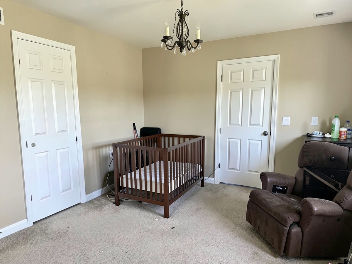 beige nursery features two white doors, a brown crib, and a brown recliner, with a black dresser and chandelier adding contrast.