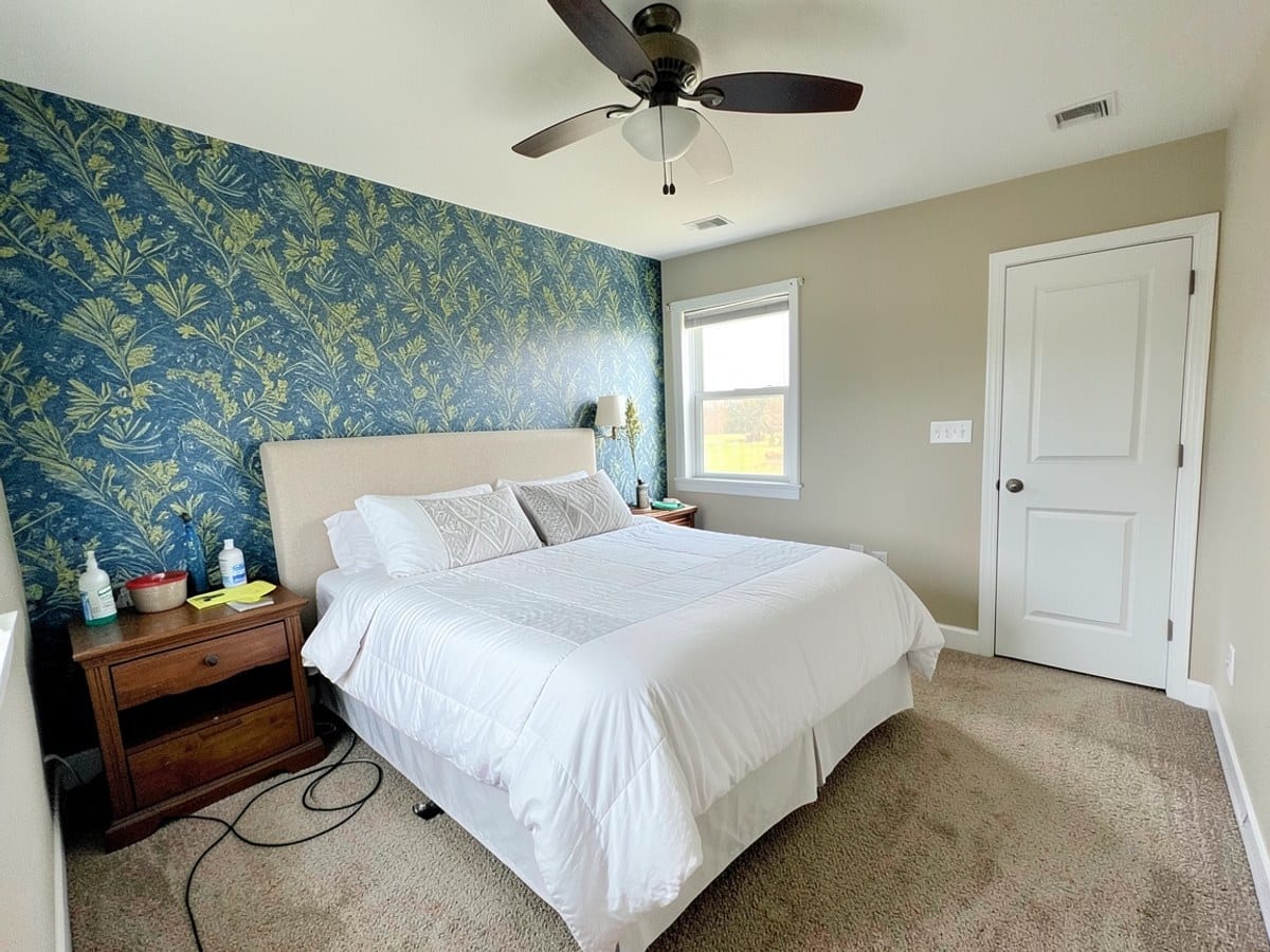 queen-sized bed with white comforter and pillows sits against a blue and green patterned wall, accompanied by a nightstand, window, white door, and ceiling fan in a beige-carpeted bedroom.