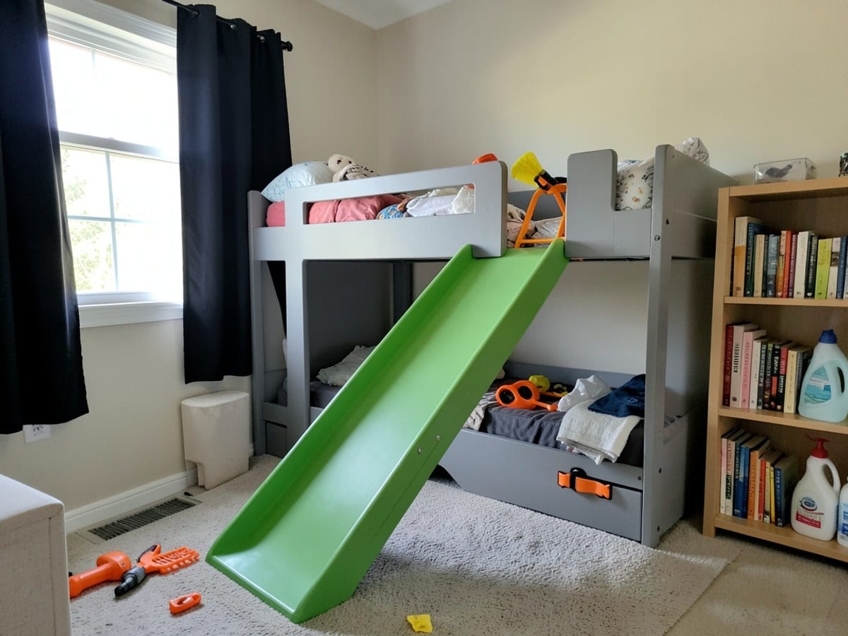 child's bedroom features a gray bunk bed with a green slideslide, orange tools, and a bookshelf, bathed in soft light from a window with black curtains.