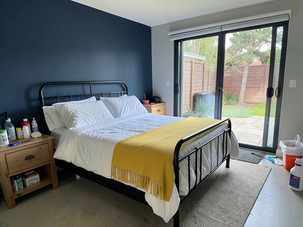 modern bedroom features a black metal bed frame, yellow throw blanket, white comforter, dark blue accent wall, wooden nightstands, sliding glass door to patio, and large window.