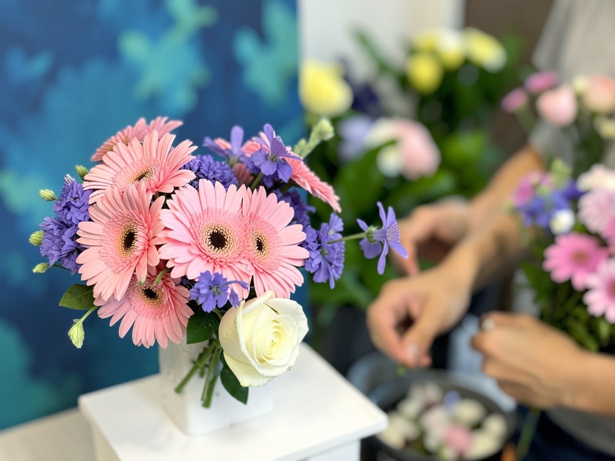 bouquet of pink gerberas, purple flowers, and a white rose, arranged on a white card, captures the viewer's attention against a blurred blue and green background.