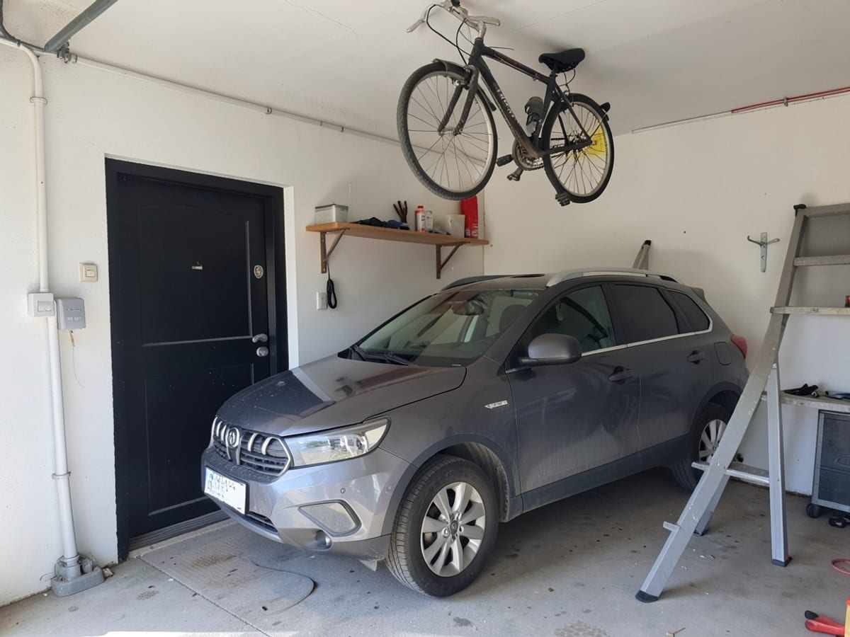 gray SUV is parked inside a white garage with a black door, a wooden shelf, and a gray ladder, while a bicycle hangs from the ceiling.