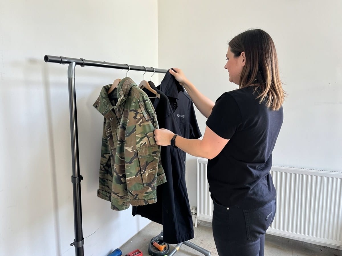 Two women examine clothing on a rack, one holding a camouflage jacket and the other a black shirt, against a white wall with a radiator.