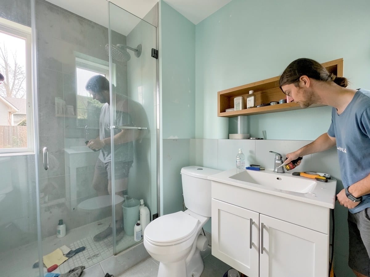 modern bathroom features a glass shower, white toilet, and sink, with a wooden shelf and white cabinet adding contrast to the light blue-green walls.