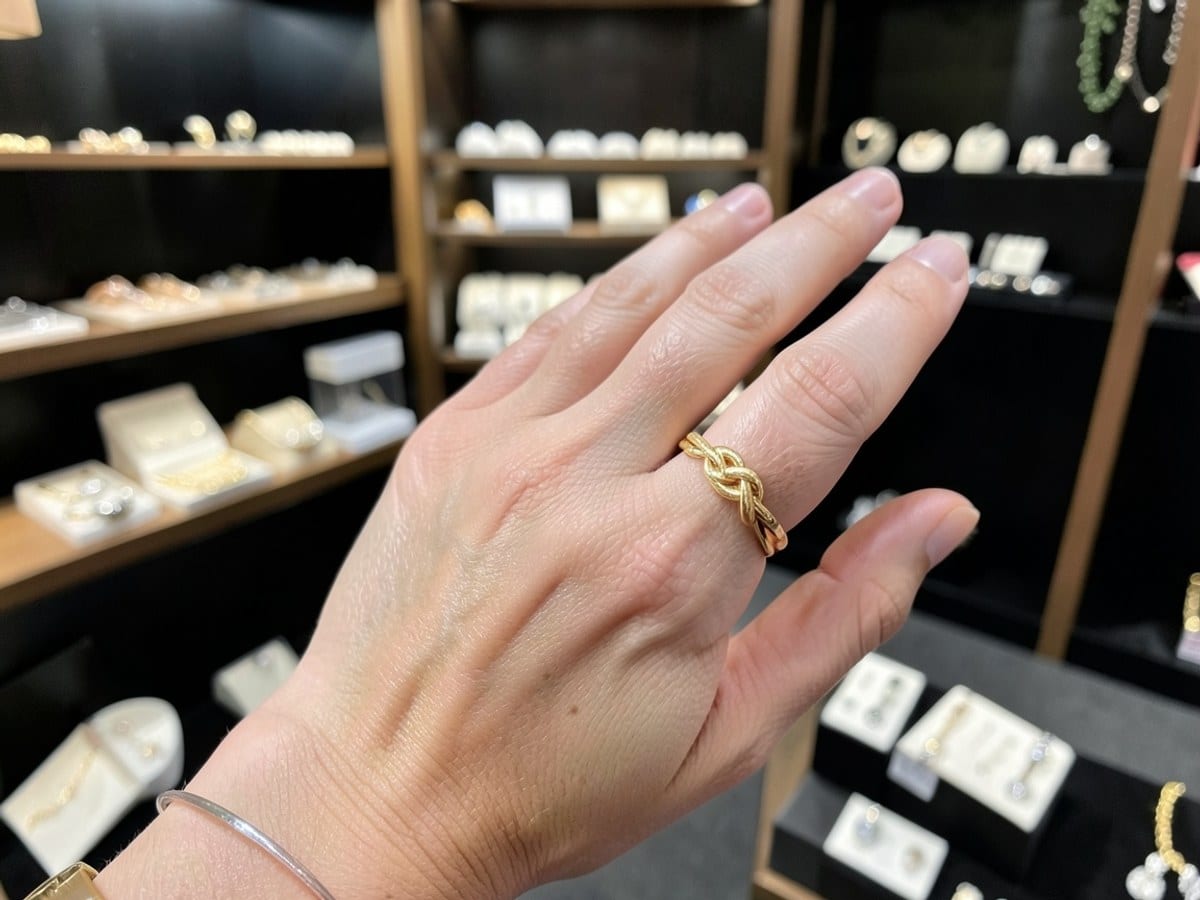 gold ring with a knot design is centered on a person's finger, with the hand's palm facing down and the index finger slightly curled, against a black background.