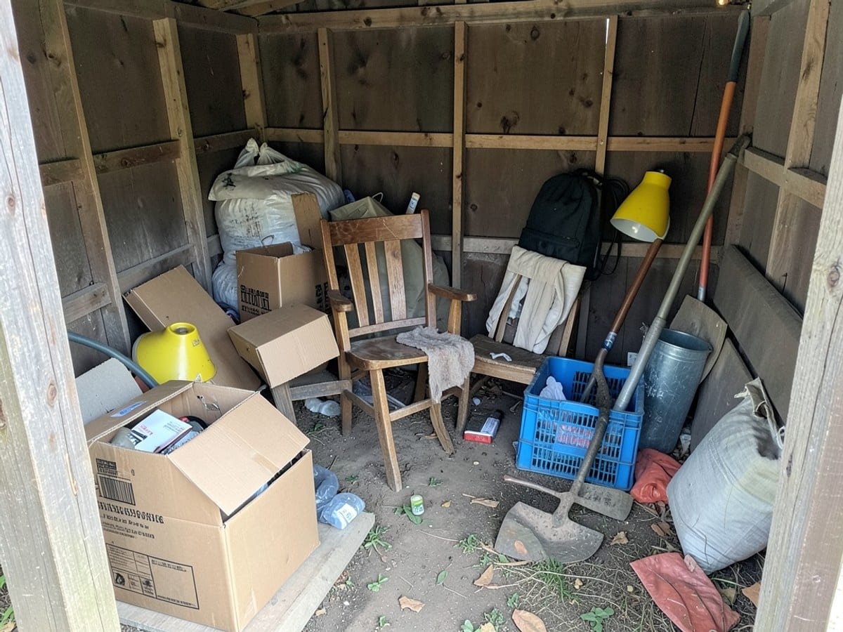 cluttered wooden shed contains various objects, including cardboard boxes, a wooden chair, a yellow lamp, a blue crate, and a gardening tool.
