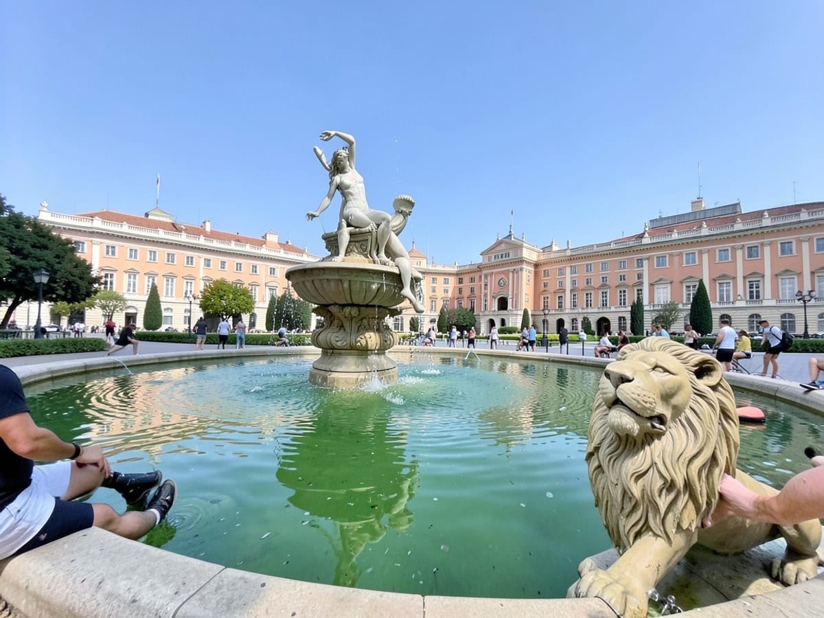 Charisma Park in Madrid, Spain, features a fountain with a woman on a throne and a lion statue, framed by the Royal Palace and the Alhambra, with a blue sky and green water reflecting the park's archi