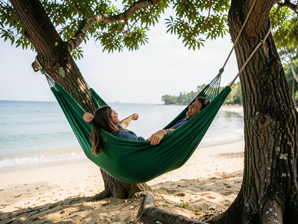 woman relaxes in a green hammock, suspended between two trees, overlooking a sandy beach and calm, reflective water.