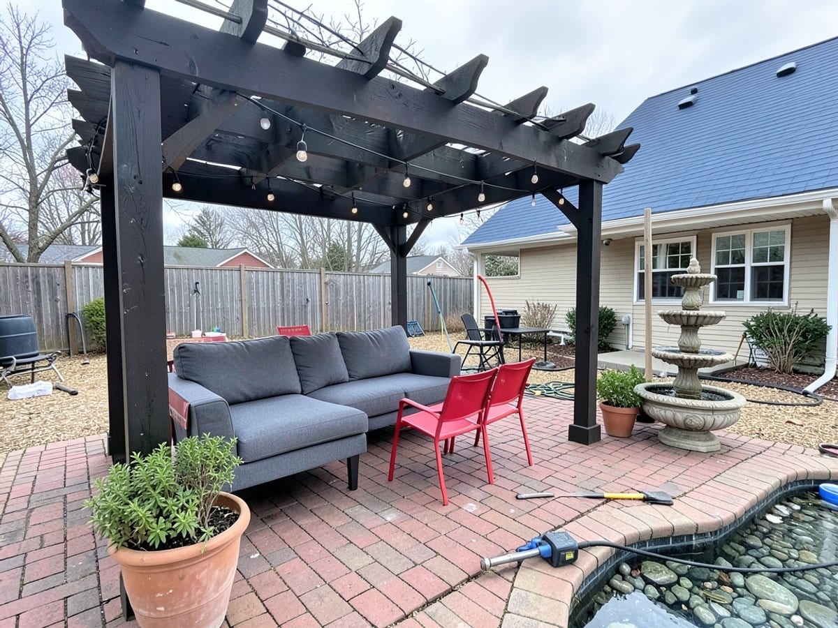dark wooden pergola with string lights shelters a gray sofa and red chairs in a brick patio, accompanied by a stone fountain, potted plants, and a house with a blue roof.